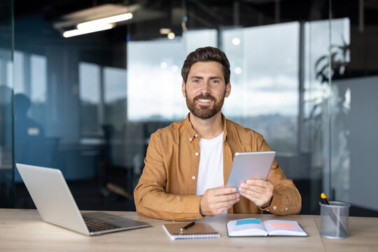 A professional man sits at a desk in an office, holding a tablet and smiling, with a laptop and open notebook on the desk. - Powered by Adobe