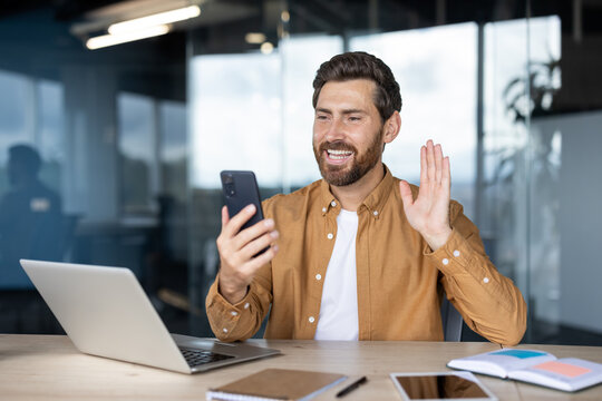 Bearded professional waving during a video call working on a smartphone and laptop in a bright, modern office. Interaction represents remote communication, technology, and professional networking.