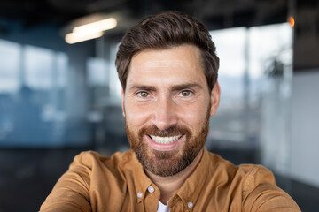 Obraz premium A cheerful man, with a bright smile, takes a selfie in a contemporary office setting. This image reflects positivity, confidence, and warmth, ideal for business and lifestyle related visuals.
