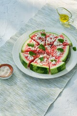 Appetizer, watermelon chunks with rind, sprinkled with crumbled feta cheese, drizzled with olive oil and garnished with mint leaves on a ceramic plate.