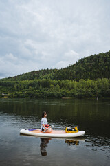 A woman paddleboards gracefully on a serene lake, surrounded by vibrant hills and cloudy skies,...
