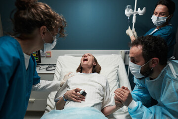 Caucasian young adult woman lying in hospital bed experiencing labor pain surrounded by medical...