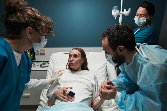 Caucasian young adult woman lying in hospital bed giving birth assisted by two female medical professionals while man holding her hand supporting her - Powered by Adobe