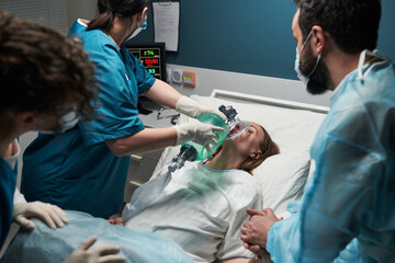 Caucasian young adult woman lying in hospital bed receiving oxygen from female medical worker while two healthcare professionals standing nearby monitoring patient condition