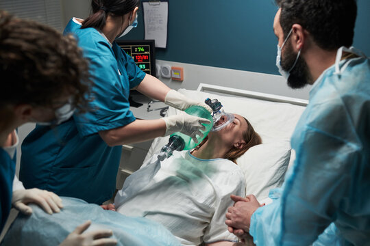 Caucasian young adult woman lying in hospital bed receiving oxygen from female medical worker while two healthcare professionals standing nearby monitoring patient condition
