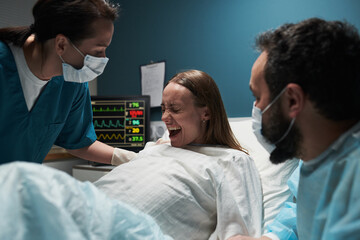 Caucasian young adult woman lying in hospital bed experiencing labor pain, assisted by female nurse...