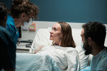 Caucasian young adult woman lying in hospital bed looking at female doctor wearing mask while male...