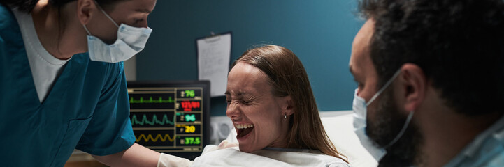 Caucasian young adult woman experiencing labor pain while lying in hospital bed, assisted by female...