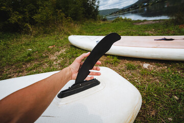 A person holds a sleek black fin, preparing to install it on a white paddleboard by a tranquil lake, surrounded by lush greenery, ideal for themes of outdoor adventure and exploration