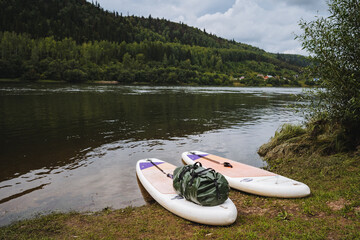 Two colorful paddleboards rest by a scenic riverbank, surrounded by greenery. This picturesque spot is perfect for summer water sports and outdoor fun, offering peace and memorable moments
