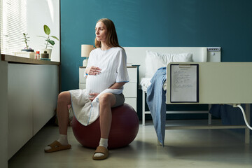 Caucasian pregnant young adult woman sitting on exercise ball in hospital room holding belly,...