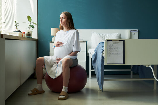 Caucasian pregnant young adult woman sitting on exercise ball in hospital room holding belly, gazing out window, medical chart visible on bed, preparing for childbirth - Powered by Adobe