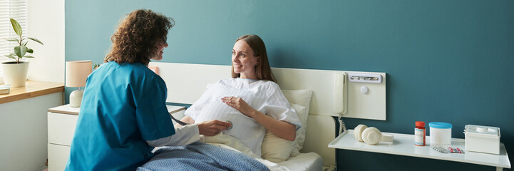 Caucasian pregnant young adult woman lying in hospital bed smiling at female nurse performing medical checkup, nurse holding stethoscope, medical equipment on bedside table