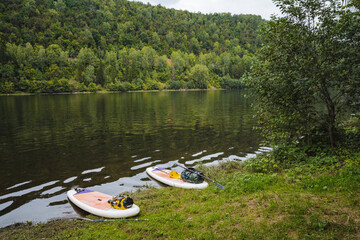 A tranquil riverside view with paddleboards at the shore and vibrant green trees reflecting in calm water, creating an ideal setting for outdoor adventures and relaxation in nature
