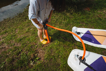 A person uses a hand pump to inflate two standup paddleboards on lush grass by a tranquil water body, preparing for a fun day of adventure, great memories, and enjoying natures beauty