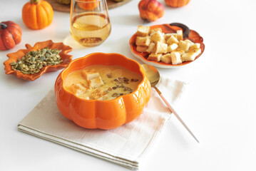 Creamy pumpkin soup with croutons and seeds, served in a pumpkin-shaped bowl on white wooden table.
