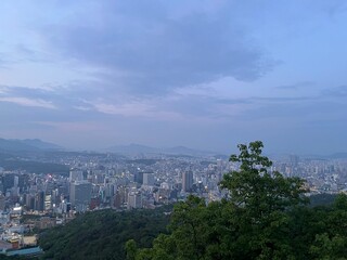 Panoramic view of Seoul city skyline at dusk from mountain viewpoint, vibrant urban lights with mountains in the background under a soft blue evening sky