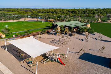 A playground with wooden structures and children's slides sits on the sand. In the background, a football field covered in lush grass, a vast forest area, and mountains on the horizon. Kundu, Belek
