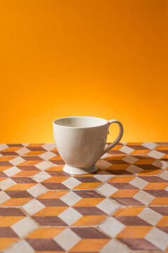 Empty white teacup on a checker pattern tile by a glowing orange wall.