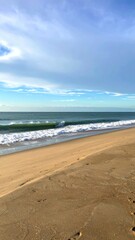 Fototapeta premium Gentle waves roll onto a sandy beach under a blue sky with light clouds.