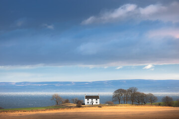 A lone white house sits on golden farmland overlooking the choppy sea and horizon of the Firth of Forth between Kirkcaldy and Kinghorn in rural Fife, Scotland.