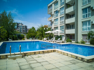 Clean outdoor pool with clear blue water, sun loungers, and umbrellas near modern apartment building. Peaceful summer vibe and relaxation area.