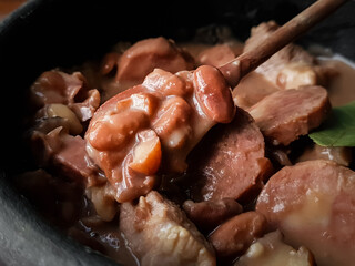 Closed up of a spoon scooping feijoada, a typical Brazilian dish with black beans, bacon, sausage, and pork in a clay pot