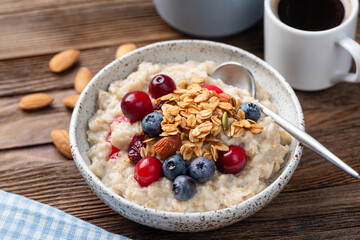 Oatmeal porridge with berries and crunchy granola topping, served with cup of coffee, wooden table background. Healthy breakfast meal