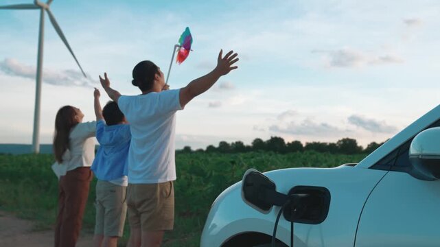 Concept of progressive happy family enjoying their time at wind farm with electric vehicle. Electric vehicle driven by clean renewable energy from wind turbine generator for charging station.