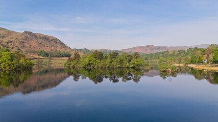 The tranquil lake reflects the surrounding hills and lush green islands of Rydal Water in Lake District England beautifully.