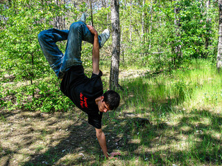 A young guy in a black T-shirt performs an acrobatic stunt standing on one hand in a green forest	
