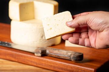 Semi-cured cheese made in Minas Gerais, Brazil, with a human hand holding a slice, on a rustic surface and dark background, selective focus.