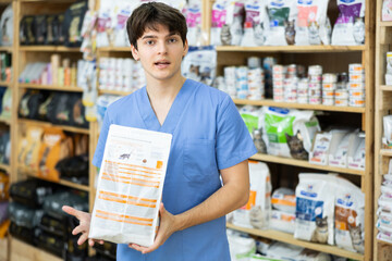 Friendly young male doctor of veterinary medicine is standing in pet store with package of food in hands. Qualified assistance in selection of food for tamed animals according to disease.