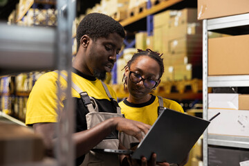 Employees team examining package data on laptop for order fulfillment in distribution hub, verifying pallets tracking numbers for shipment protocols. Efficient e-commerce facility.