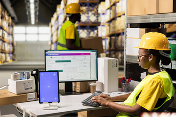 Black woman typing awb tracking info next to isolated mockup display, overseeing shipment details with hi vis vest and helmet in warehouse setting. Monitoring package tracking on software.