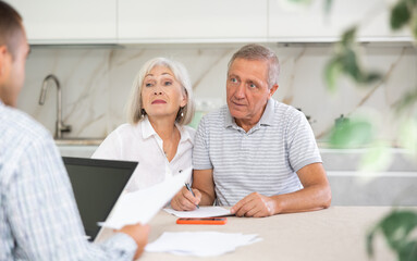 Young salesman explaining terms of deal to elderly couple sitting in kitchen. Health insurance concept