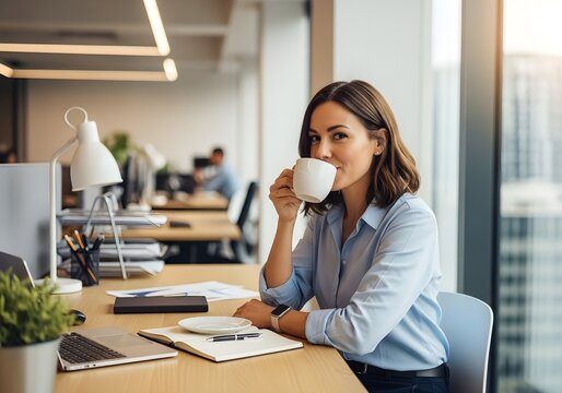Professional Woman Enjoying Coffee Break at Desk in Modern Office with Laptop, Notebook and Office Supplies Scattered About