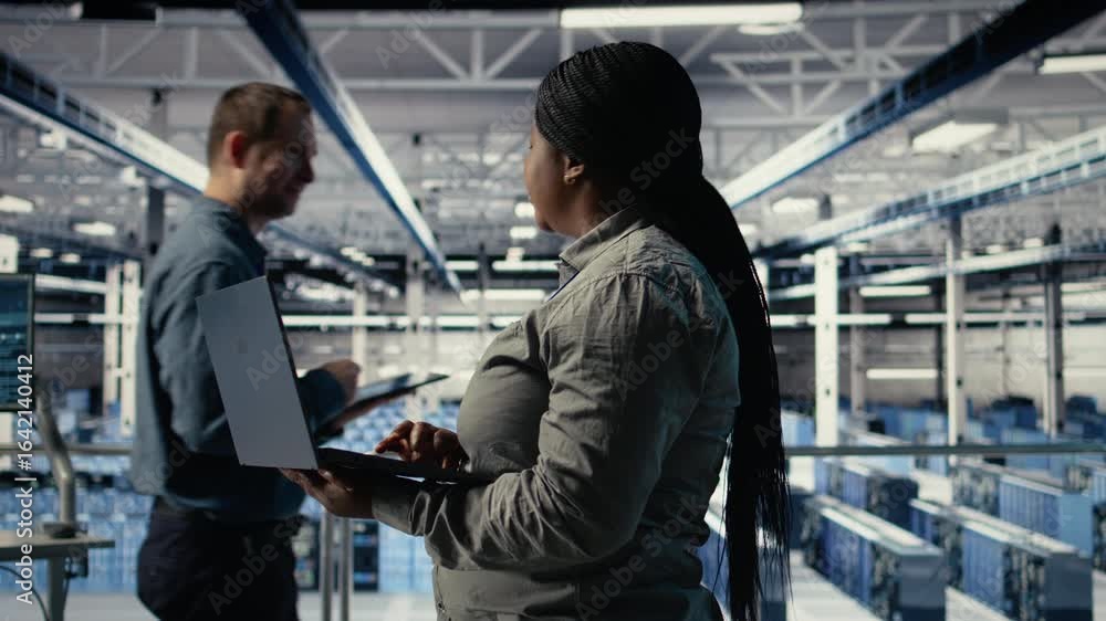 Poster it expert monitors server farm activity using laptop while walking in facility. african american wom