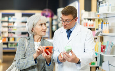 Concerned old woman showing to middle-aged male pharmacist QR-code of product on her smartphone in chemist's shop