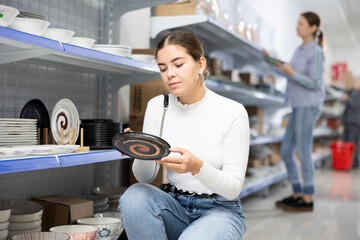 Young woman in supermarket is choosing plates. Female buyer examines ceramic kitchen utensils with...