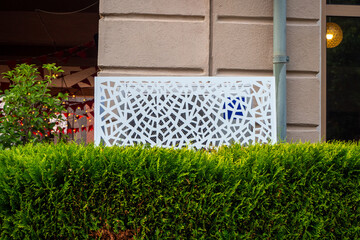 Protective decorative metal grate and trimmed bushes cover the air conditioning unit near building wall. Improvement of courtyard area. Real photo