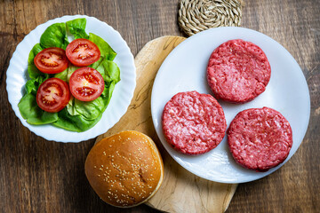 Three raw beef burger patties on a plate next to a bun, lettuce, and sliced tomatoes on a wooden surface.