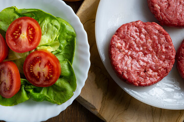 Three raw beef burger patties on a plate next to a bun, lettuce, and sliced tomatoes on a wooden surface.