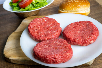 Three raw beef burger patties on a plate next to a bun, lettuce, and sliced tomatoes on a wooden surface.