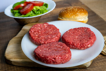 Three raw beef burger patties on a plate next to a bun, lettuce, and sliced tomatoes on a wooden surface.