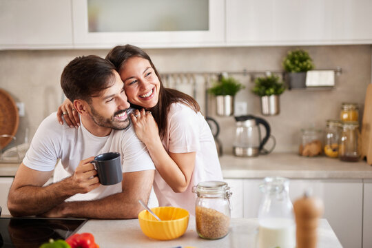 A couple shares joyful moments in a bright kitchen while preparing breakfast. The man holds a coffee cup, and the woman hugs him, smiling happily amidst various ingredients.