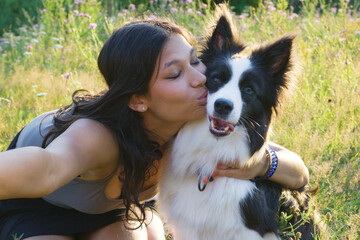 Indian woman kissing her Border Collie dog while taking a selfie in a sunny meadow, showing love...