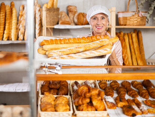 Senior female baker sells baguettes, shows many ready products. Authors bakery, fresh pastry. In...