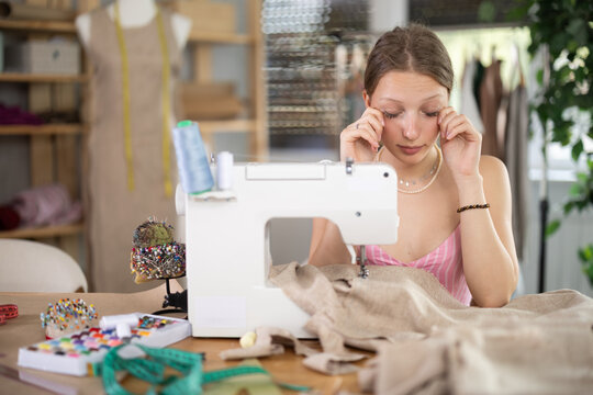 Young female seamstress with tired eyes sewing clothes on sewing machine in workshop