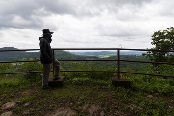 Scenic viewpoint at Phra That Phu Phan Forest Monastery during rainy season, lush green mountains with a male tourist standing and enjoying the panoramic view.